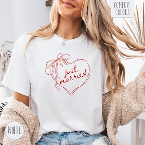 Op de afbeelding: Wit T-shirt met een rood hartvormig ontwerp en de woorden "just married" in cursief. Het hart is versierd met een strik en linten. Op het shirt staan ook de woorden "Comfort Colors" en "White".