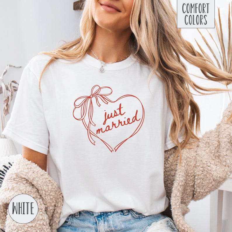 Op de afbeelding: Wit T-shirt met een rood hartvormig ontwerp en de woorden "just married" in cursief. Het hart is versierd met een strik en linten. Op het shirt staan ook de woorden "Comfort Colors" en "White".