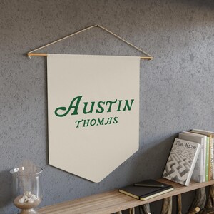 May include: A beige canvas banner with a wooden dowel at the top. The banner has the text "Austin Thomas" in green cursive lettering.