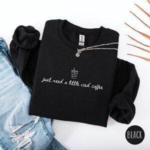 K&ouml;nnte beinhalten: Schwarzes Sweatshirt mit dem wei&szlig;en Schriftzug "just need a little iced coffee" und einer kleinen Eiskaffee-Grafik. Eine silberne Halskette mit Anh&auml;nger liegt auf dem Sweatshirt. Das Wort "BLACK" steht in einem schwarzen Kreis.