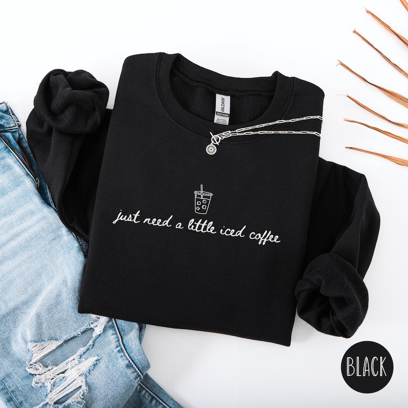 K&ouml;nnte beinhalten: Schwarzes Sweatshirt mit dem wei&szlig;en Schriftzug "just need a little iced coffee" und einer kleinen Eiskaffee-Grafik. Eine silberne Halskette mit Anh&auml;nger liegt auf dem Sweatshirt. Das Wort "BLACK" steht in einem schwarzen Kreis.