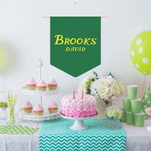 May include: A green banner with the text "BROOKS DAVID" in yellow letters hangs above a table with a two-tiered cake stand holding pink and white cupcakes. A pink and white cake with pink frosting sits on a white cake stand. There are green and white polka dot cups and a green and white polka dot tablecloth.