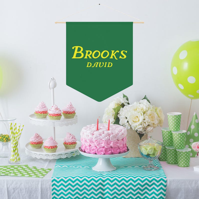 May include: A green banner with the text "BROOKS DAVID" in yellow letters hangs above a table with a two-tiered cake stand holding pink and white cupcakes. A pink and white cake with pink frosting sits on a white cake stand. There are green and white polka dot cups and a green and white polka dot tablecloth.