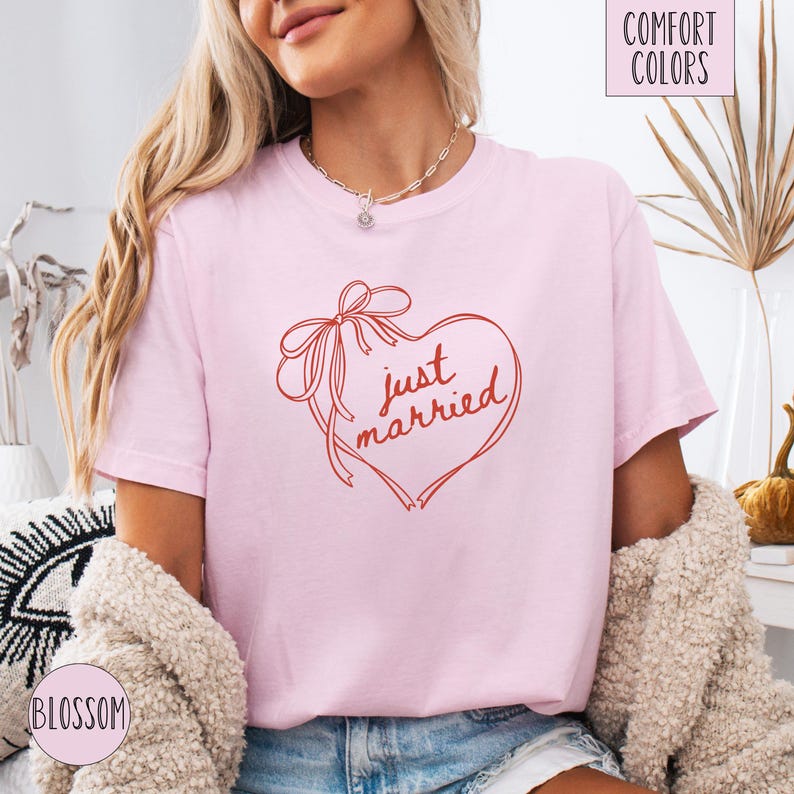 Op de afbeelding: Lichtroze T-shirt met een rood hartvormig ontwerp en de tekst "just married" in een handgeschreven lettertype. Het hart heeft een strik. Onderdeel van de "Comfort Colors" collectie.