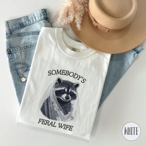 May include: White t-shirt with a black and white illustration of a raccoon wearing a bridal veil. The text "Somebody's Feral Wife" is printed below the illustration.