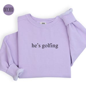 May include: A lavender sweatshirt with the text "he's golfing" printed on the front.