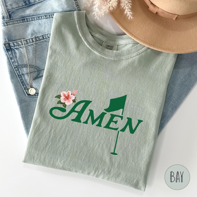 May include: A light green t-shirt with a green golf flag and the word "AMEN" in green script. A pink flower with green leaves is above the word "AMEN".
