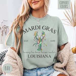 May include: A light green t-shirt with a black graphic of a lobster with gold and green accents. The text "Mardi Gras" is above the lobster, and the text "Est. 1699" is below the lobster. The text "Laissez les bons temps rouler Louisiana" is below the lobster.