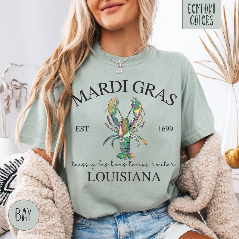 May include: A light green t-shirt with a black graphic of a lobster with gold and green accents. The text "Mardi Gras" is above the lobster, and the text "Est. 1699" is below the lobster. The text "Laissez les bons temps rouler Louisiana" is below the lobster.