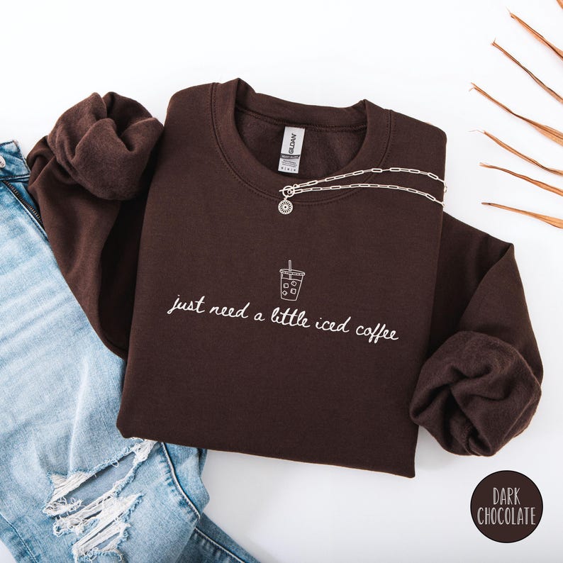 K&ouml;nnte beinhalten: Ein dunkelbraunes Sweatshirt mit dem wei&szlig;en Schriftzug "just need a little iced coffee". Eine silberne Halskette mit Anh&auml;nger liegt &uuml;ber der Vorderseite. Das Sweatshirt ist neben einer Jeans gefaltet.