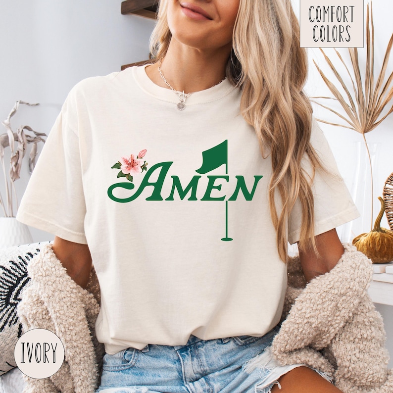 May include: A white t-shirt with a green golf flag and the word "AMEN" in green text. The shirt also has a pink flower graphic.