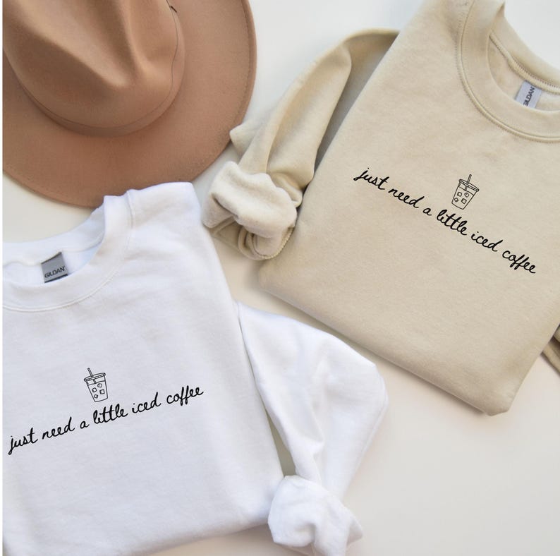 K&ouml;nnte beinhalten: Zwei Sweatshirts, eines wei&szlig; und eines beige, mit dem Text "just need a little iced coffee" in einer handschriftlichen Schriftart. Jedes Sweatshirt hat eine kleine Grafik einer Eiskaffeetasse. Ein beiger Hut ist im Hintergrund zu sehen.