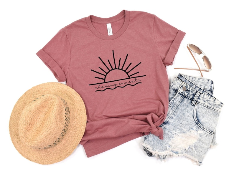 Peut inclure: Un t-shirt mauve avec un graphique noir d'un soleil couchant sur l'oc&eacute;an et le texte "chasing sunsets". Le t-shirt est associ&eacute; &agrave; un short en jean, un chapeau de paille et des lunettes de soleil.