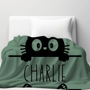 May include: A green fleece blanket with a black cat peeking over the top. The cat has large eyes and a cute expression. The blanket has the name "CHARLIE" printed in black letters.