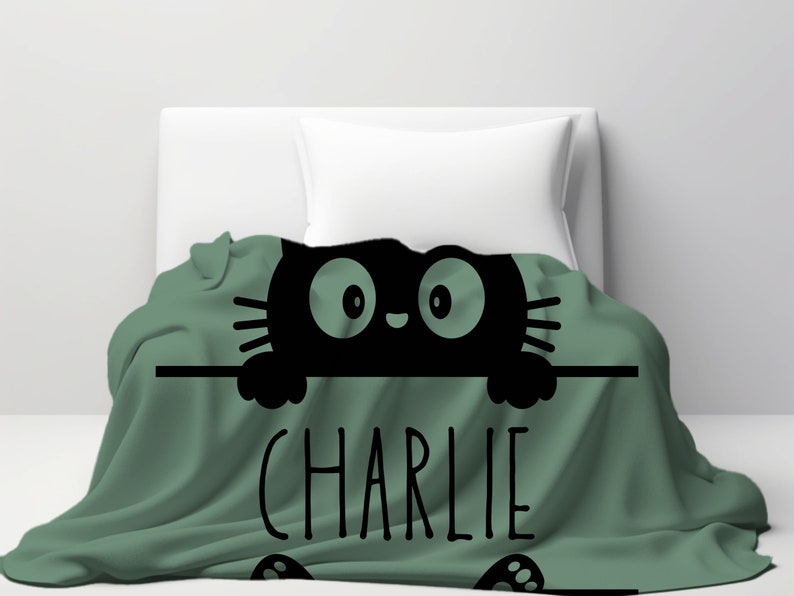 May include: A green fleece blanket with a black cat peeking over the top. The cat has large eyes and a cute expression. The blanket has the name "CHARLIE" printed in black letters.