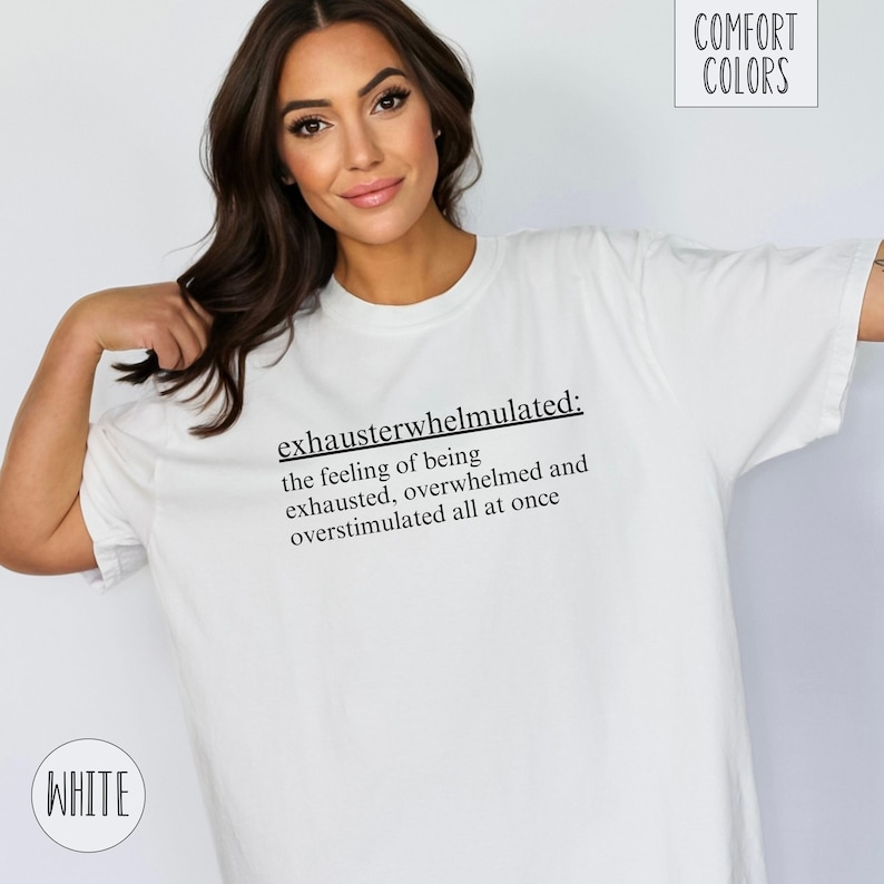 Op de afbeelding: Wit T-shirt met zwarte tekst die "exhausterwhelmulated: the feeling of being exhausted, overwhelmed and overstimulated all at once" luidt