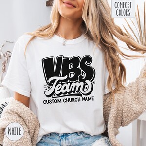 Op de afbeelding: Wit T-shirt met een zwart-witte afbeelding met de tekst "VBS Team" en "Custom Church Name".