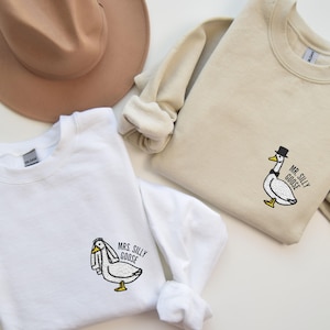 May include: Two white sweatshirts with embroidered images of geese. One sweatshirt says "Mrs. Silly Goose" and the other says "Mr. Silly Goose".