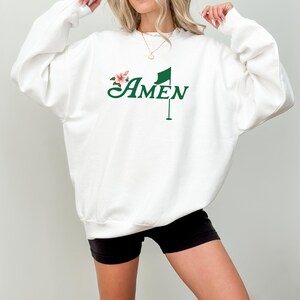 May include: White sweatshirt with a green golf flag and the word "AMEN" in a cursive font. A small pink flower is next to the flag.