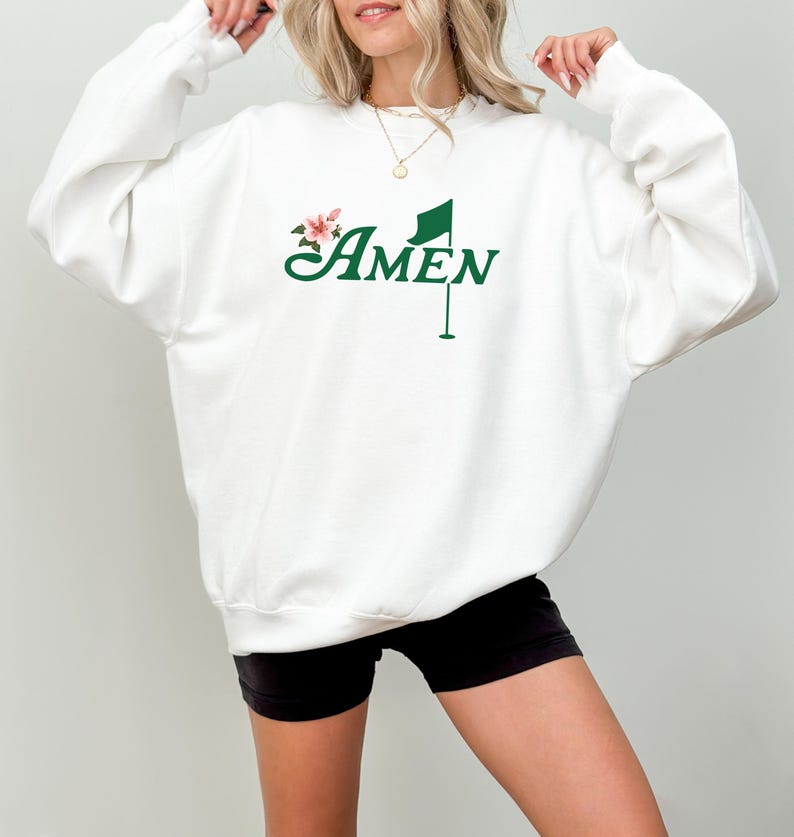 May include: White sweatshirt with a green golf flag and the word "AMEN" in a cursive font. A small pink flower is next to the flag.