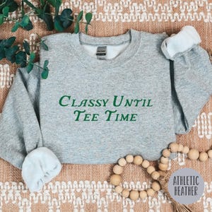 以下が含まれることがあります： グレーのトレーナーに、緑色の文字で「Classy Until Tee Time」と書いてあります。トレーナーは、茶色と白の模様のある表面の上に置かれています。