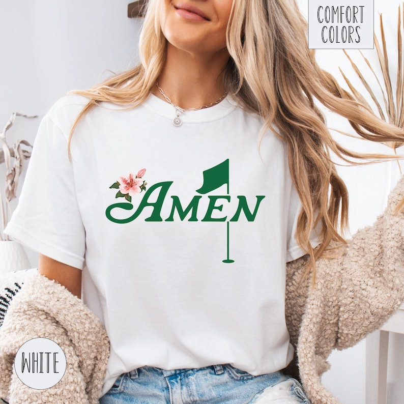 May include: White t-shirt with a green golf flag and the word "AMEN" in a green cursive font. A pink flower is behind the word "AMEN".