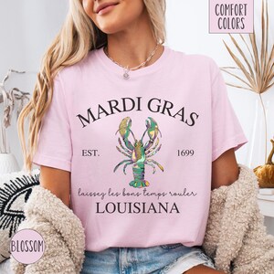 May include: Pink t-shirt with a Mardi Gras design featuring a green, gold, and purple lobster with the text "Mardi Gras", "Est. 1699", "laissez les bons temps rouler", and "Louisiana".