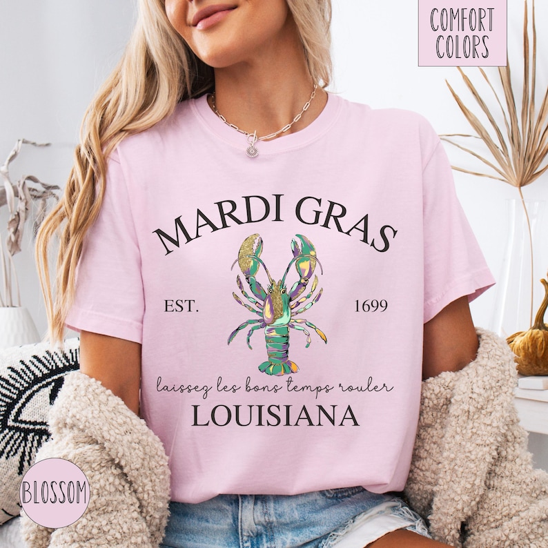 May include: Pink t-shirt with a Mardi Gras design featuring a green, gold, and purple lobster with the text "Mardi Gras", "Est. 1699", "laissez les bons temps rouler", and "Louisiana".