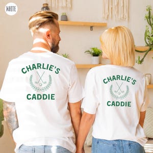 May include: Two white t-shirts with a green design on the back. The design features a golf club and ball with a wreath and the text "Charlie's Caddie".