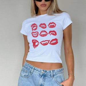 May include: White t-shirt with a crew neck featuring a red graphic of various vampire mouth designs. The shirt is short-sleeved and worn with light blue denim jeans.