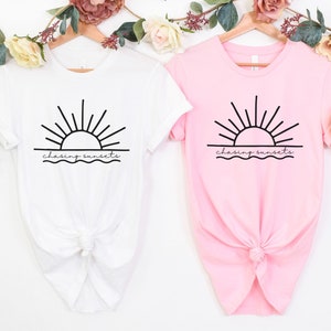 Peut inclure: Deux t-shirts avec un motif de soleil couchant sur l'oc&eacute;an et le texte "chasing sunsets". Un t-shirt est blanc et l'autre est rose.