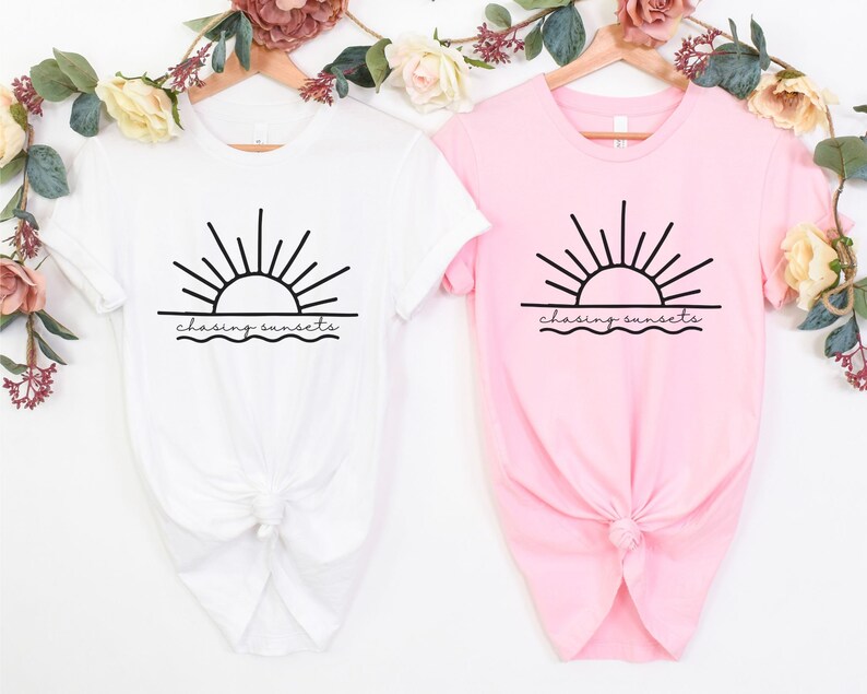 Peut inclure: Deux t-shirts avec un motif de soleil couchant sur l'oc&eacute;an et le texte "chasing sunsets". Un t-shirt est blanc et l'autre est rose.