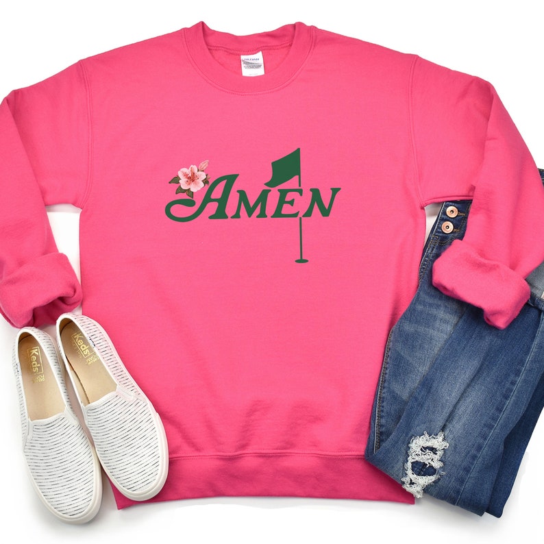 May include: Pink sweatshirt with a green golf flag and the word "AMEN" in a green cursive font. A small pink flower is above the word "AMEN".