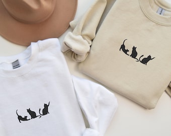 May include: Two sweatshirts with a black cat silhouette design. One sweatshirt is white and the other is beige.
