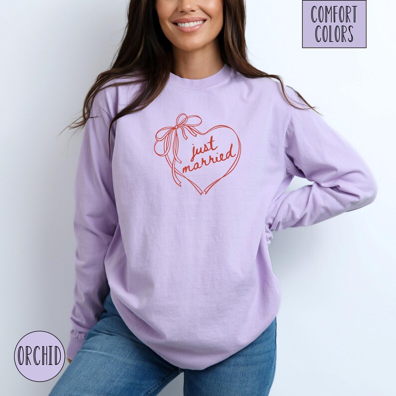 Op de afbeelding: Een lila shirt met lange mouwen met een rood hartontwerp met de tekst "just married". Het hart heeft een strik. Het shirt is voorzien van de labels "Comfort Colors" en "Orchid".