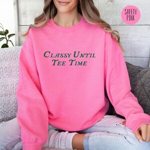 以下が含まれることがあります： 緑色の文字で「CLASSY UNTIL TEE TIME」と書かれたピンク色のスウェットシャツです。