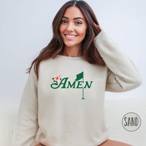 May include: A beige sweatshirt with a green golf flag and the word "AMEN" in a green cursive font. A pink flower is to the left of the word "AMEN".