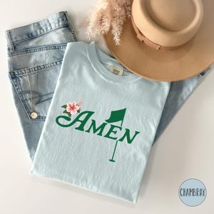 May include: A light blue t-shirt with a green golf flag and the word "AMEN" in a green cursive font. A pink flower is above the word "AMEN".