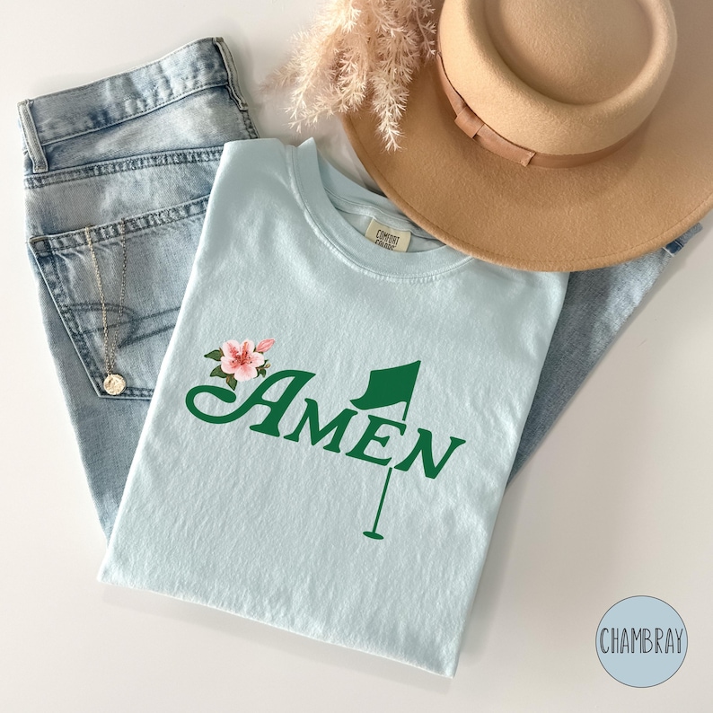May include: A light blue t-shirt with a green golf flag and the word "AMEN" in a green cursive font. A pink flower is above the word "AMEN".