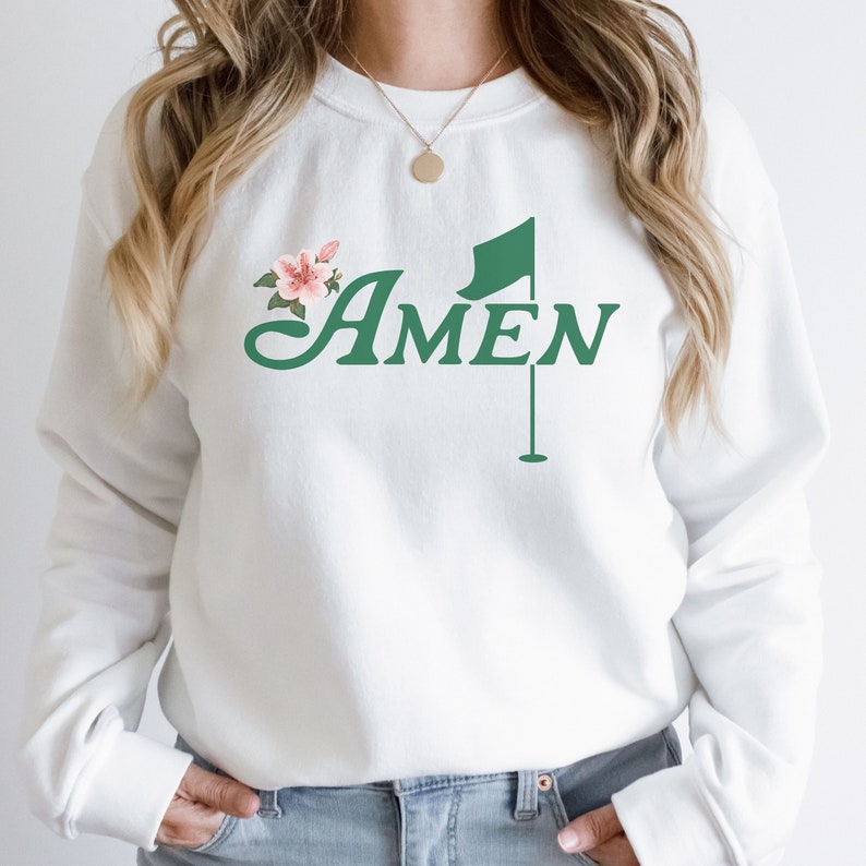 May include: White sweatshirt with a green golf flag and the word "AMEN" in a cursive font. A pink flower is to the left of the word.