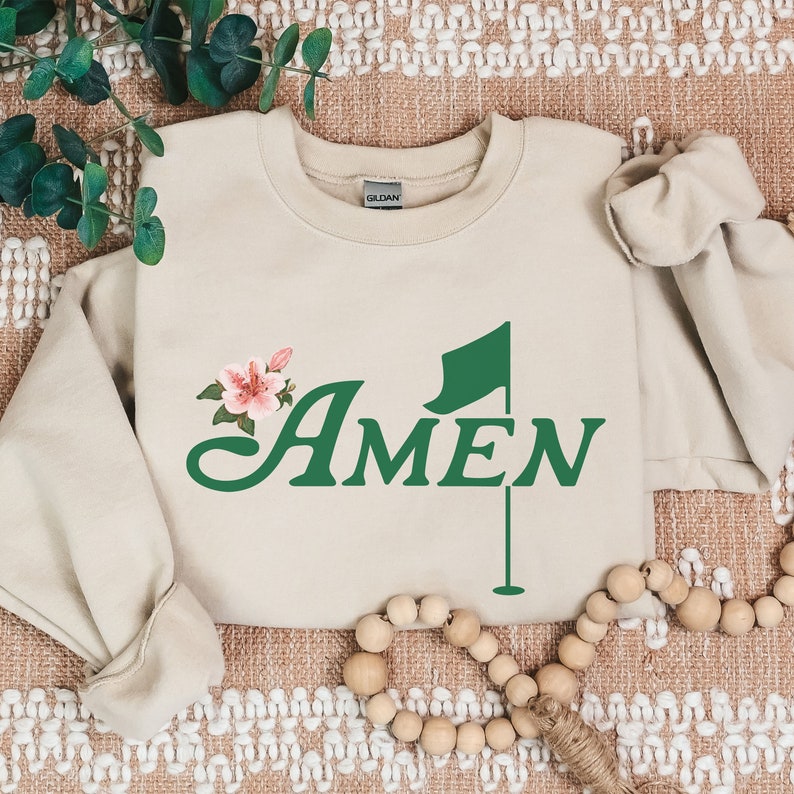 May include: A beige sweatshirt with a green golf flag and the word "AMEN" in green cursive lettering. A pink flower is behind the word "AMEN".