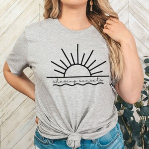 Peut inclure: Un t-shirt gris avec un graphique noir d'un soleil couchant sur l'eau. Le texte "chasing sunsets" est &eacute;crit sous le graphique.