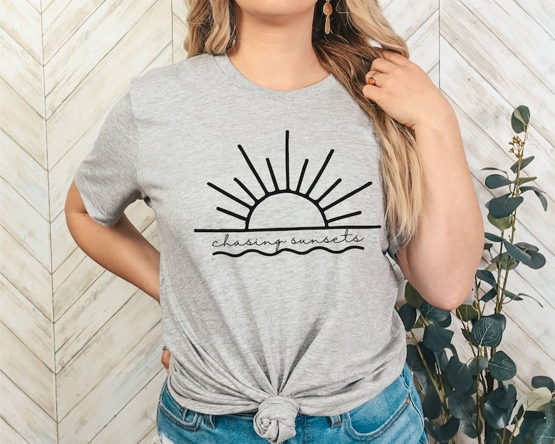 Peut inclure: Un t-shirt gris avec un graphique noir d'un soleil couchant sur l'eau. Le texte "chasing sunsets" est &eacute;crit sous le graphique.