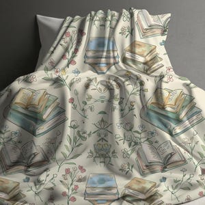 May include: A cream-colored throw blanket with a floral pattern featuring green leaves and pink, blue, and yellow flowers. The blanket has a repeating pattern of stacks of books with open pages.