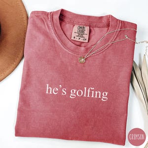 May include: A crimson red t-shirt with the text "he's golfing" in white lettering. The shirt is made of a soft, comfortable fabric and has a relaxed fit.
