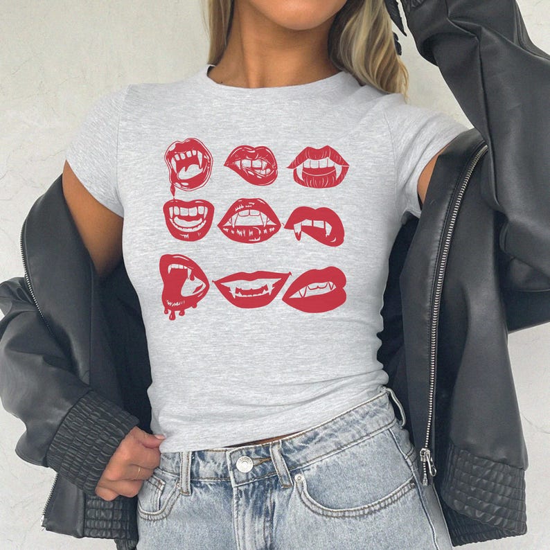 May include: A light gray t-shirt featuring a grid of nine red lip illustrations with fangs. The short-sleeved shirt is paired with blue jeans and a black leather jacket. The design has a spooky, Halloween theme.
