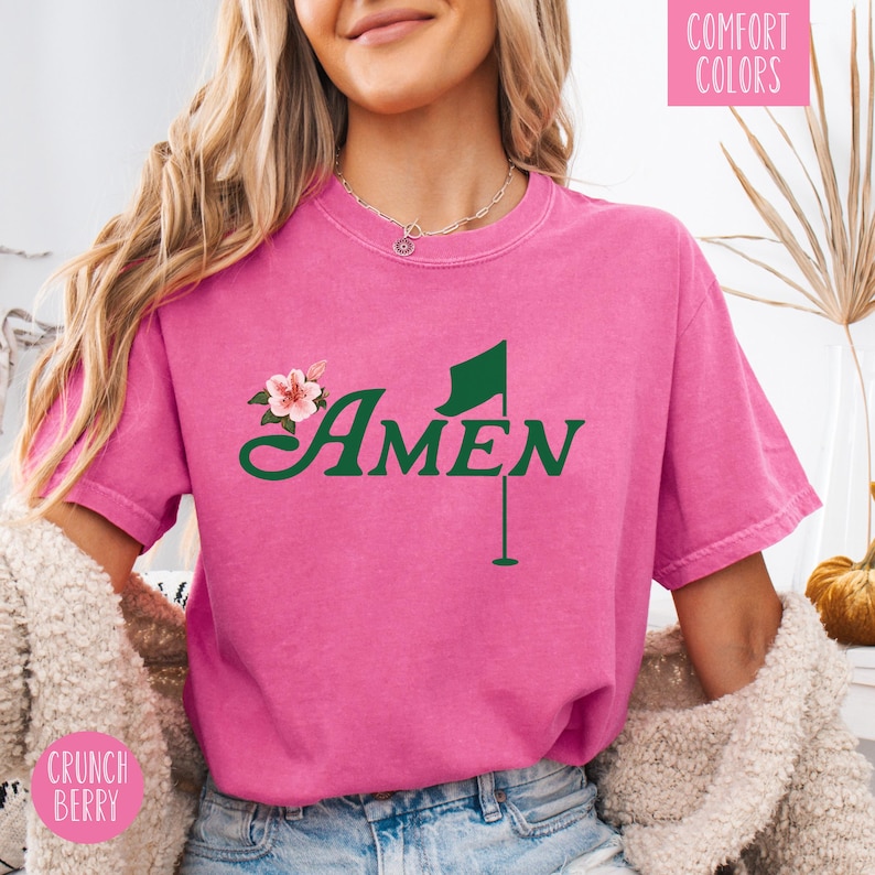May include: Pink short-sleeved t-shirt with a green golf flag and the word "AMEN" in a green cursive font. The shirt has a floral design on the left side of the chest.