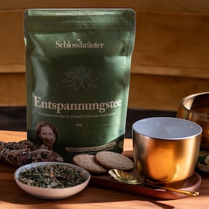 Könnte beinhalten: Ein grüner Beutel Schlosskräuter Entspannungstee, ein entspannender und schlaffördernder Kräutertee, mit einem Frauenbild auf der Vorderseite. Der Beutel liegt auf einem Holztisch mit einer goldenen Teetasse, einer Schale mit getrockneten Kräutern und einer Klangschale.