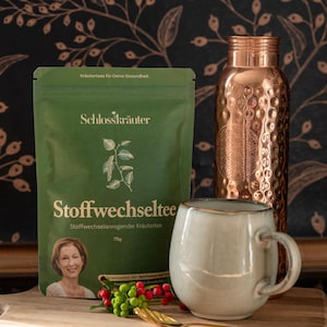 Könnte beinhalten: Ein grüner Beutel Schlosskräuter Stoffwechseltee, ein stoffwechselanregender Kräutertee, mit einer Packungsgröße von 75g. Der Beutel liegt neben einer hellgrauen Keramiktasse und einer Kupferwasserflasche.
