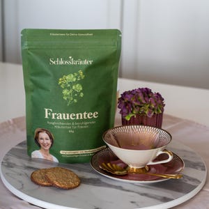May include: A green pouch of Schlosskräuter Frauentee herbal tea for women. The pouch has a picture of a woman and the text "Frauentee Ausgleichender & beruhigender Kräutertee für Frauen 65g". The pouch is sitting on a marble surface with a pink and gold teacup and saucer, two cookies, and a bouquet of purple flowers.